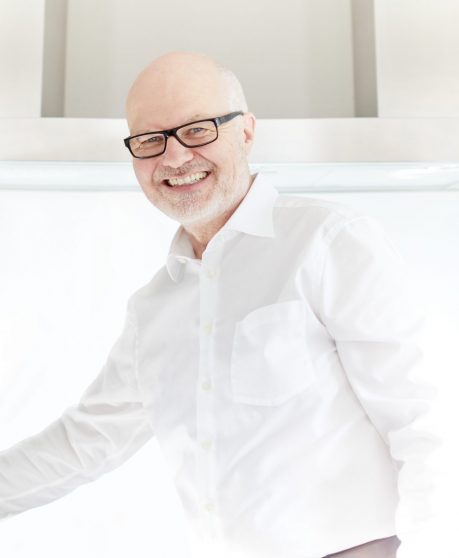Älterer Mann mit Brille und weißem Hemd lächelt freundlich in heller, moderner Umgebung.